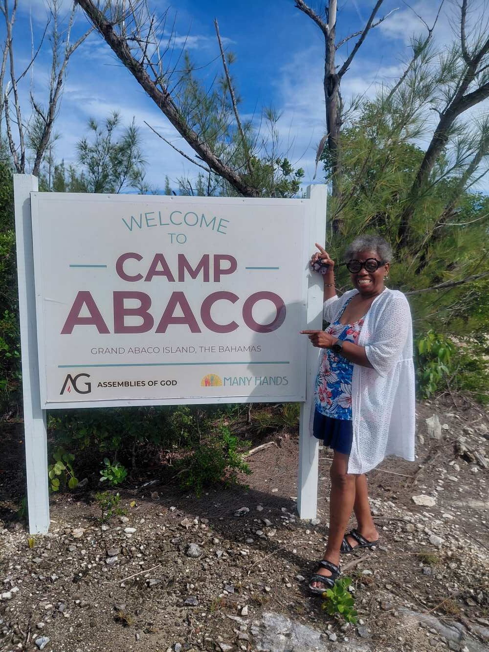A smiling volunteer points to the Many Hands logo on the Camp Abaco entrance sign during a short-term impact trip.