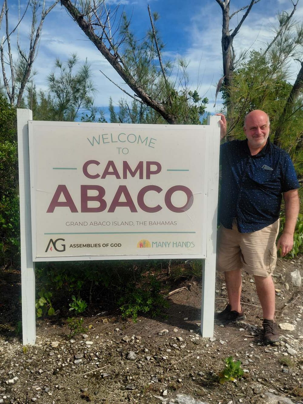 An adult volunteer stands beside the official Welcome to Camp Abaco entrance sign.
