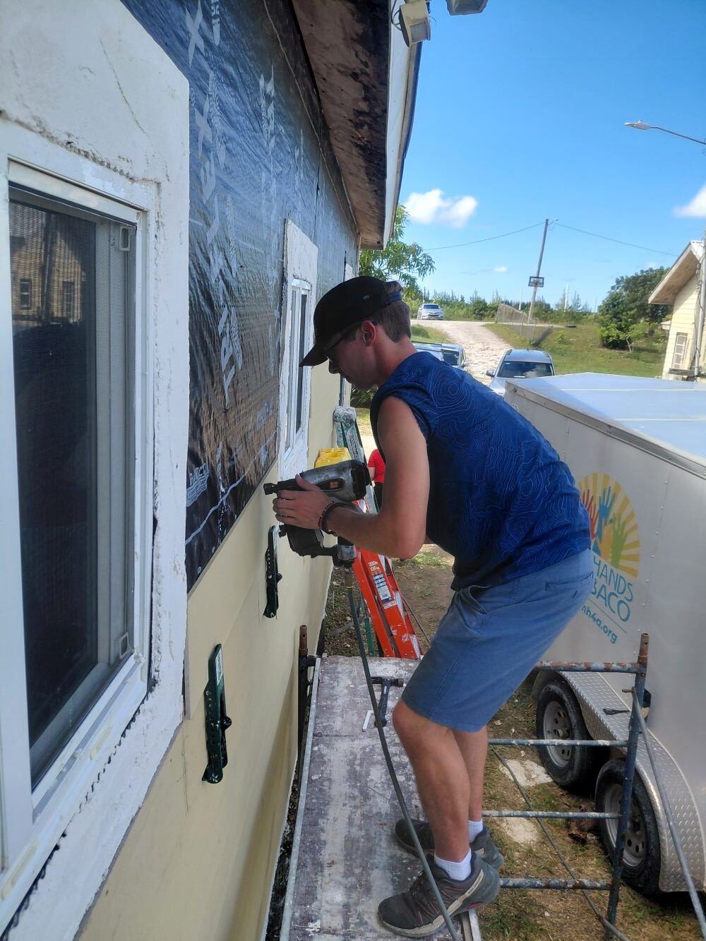 A short-term volunteer uses a pneumatic nail gun from scaffolding to secure exterior siding during a Many Hands home repair project.