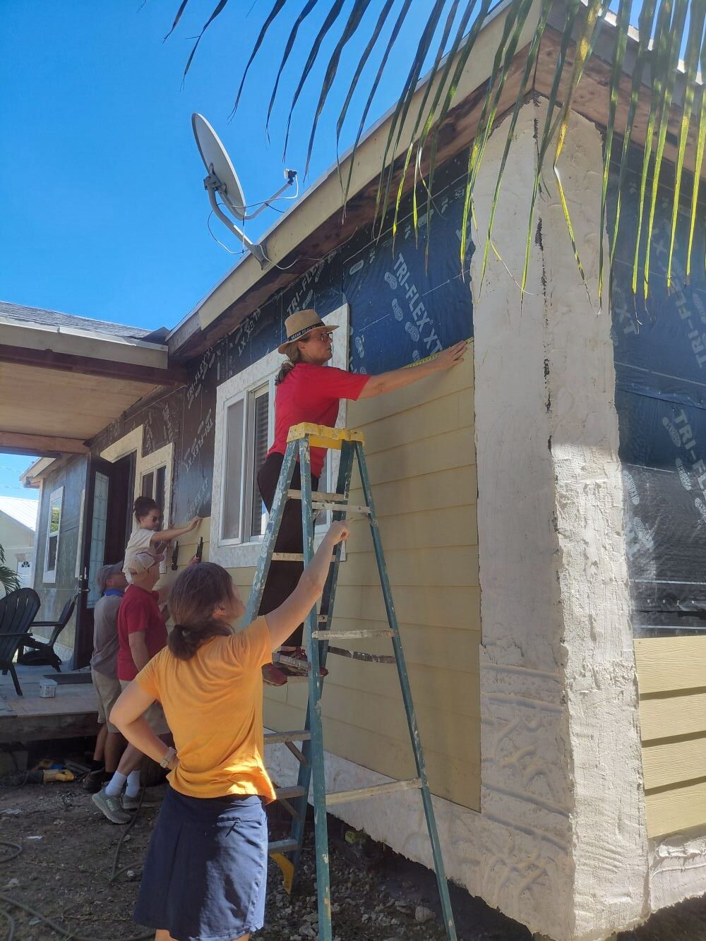 A team of volunteers collaborates on stepladders to measure and secure lap siding on a residential home restoration project.
