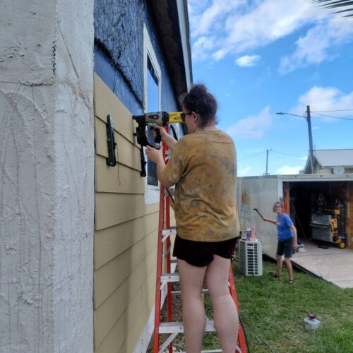 A Many Hands impact team volunteer stands on a ladder to nail lap siding onto a home, supporting the Safe Structures rebuilding program in Abaco.